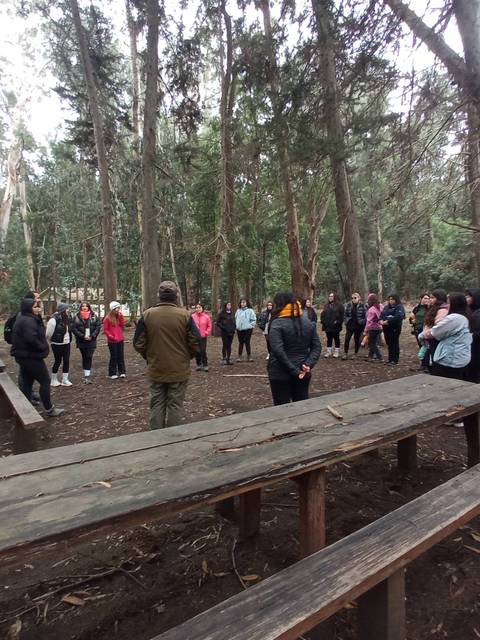 Un grupo de personas reunidas en un bosque, alrededor de mesas de madera, mientras un hombre de pie les habla.