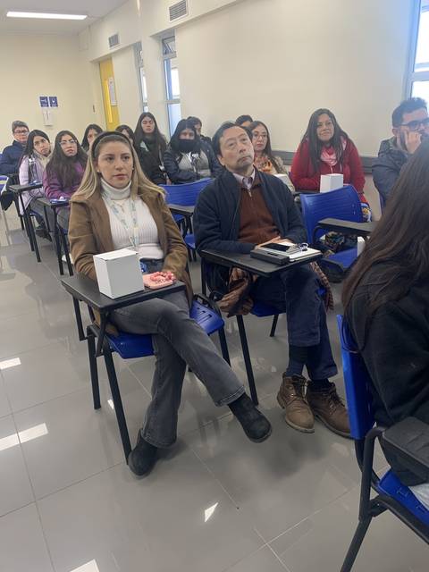 Una clase con varios estudiantes sentados en pupitres, prestando atención al frente.