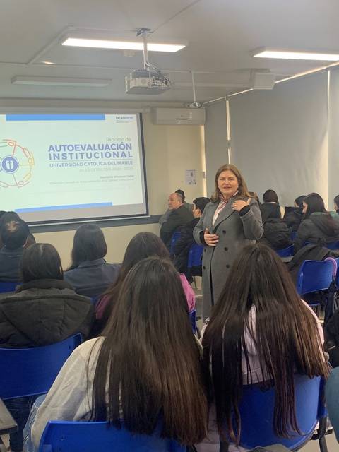 Una mujer presenta un tema sobre autoevaluación institucional frente a un grupo de estudiantes en un aula.