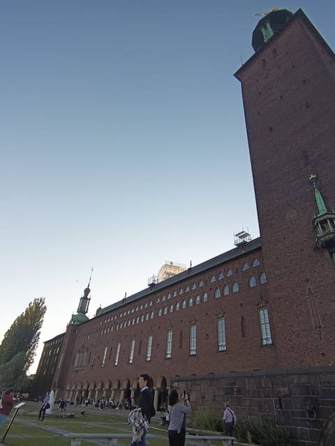 La imagen muestra un edificio de ladrillos con detalles arquitectónicos y un ambiente de encuentro al aire libre.