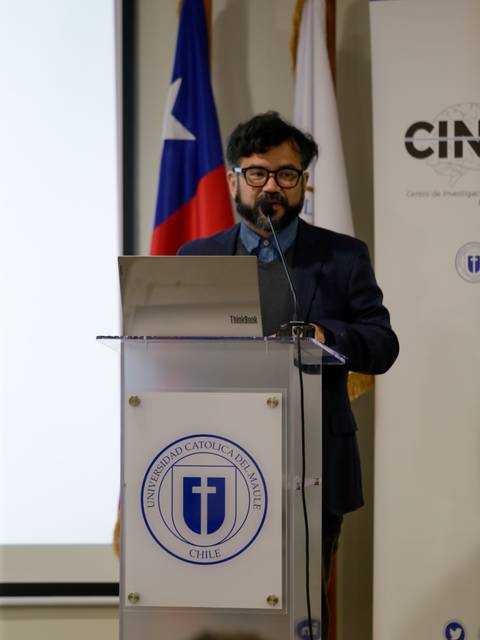 Un hombre presenta en un evento académico en una universidad, rodeado de banderas chilenas.