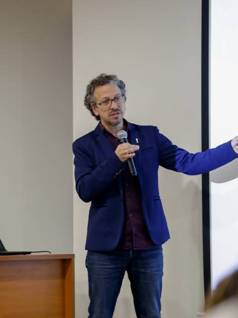 Un hombre vestido con un blazer azul está presentando en una conferencia mientras señala una pantalla con información.
