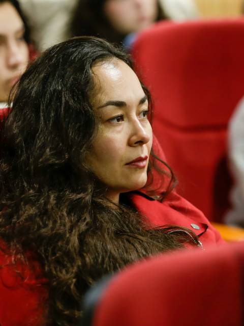 Una mujer con cabello rizado y suelto, vestida con una chaqueta roja, escucha atentamente en una sala llena de personas.
