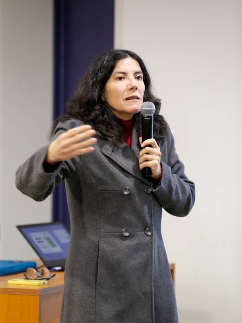 Una mujer con abrigo gris está hablando en público mientras sostiene un micrófono.