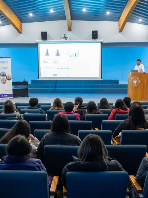Una conferencia con un orador frente a una audiencia sentada en un salón con pantallas y banderas.