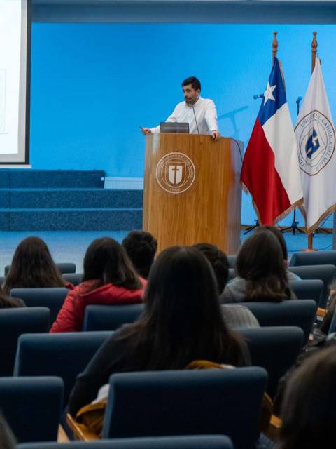 Un conferencista presenta frente a una audiencia en una sala con banderas chilenas de fondo.