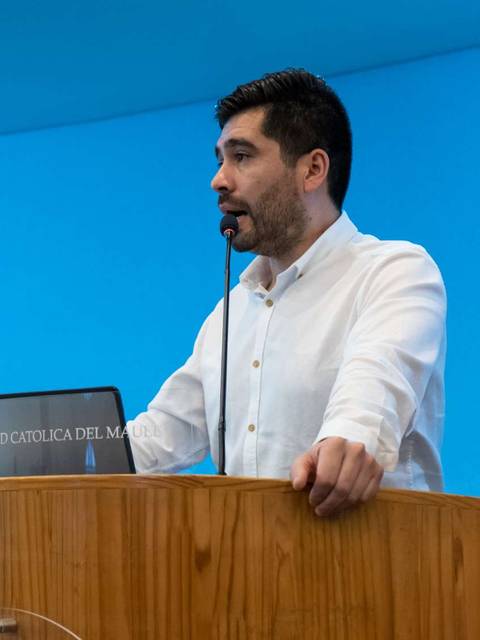 Un hombre habla frente a un podio en una sala con fondo azul y un crucifijo.