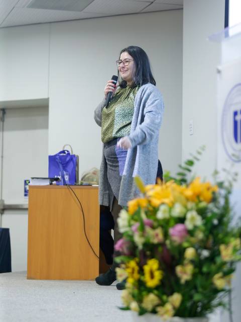 Una mujer habla en un podio, mientras sostiene un micrófono y se encuentra frente a un arreglo floral colorido.