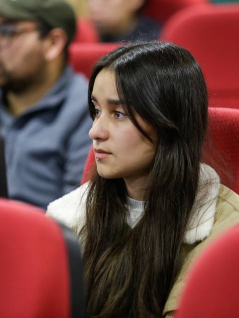 Una joven con cabello largo y oscuro escucha atentamente en un auditorio lleno de asientos rojos.