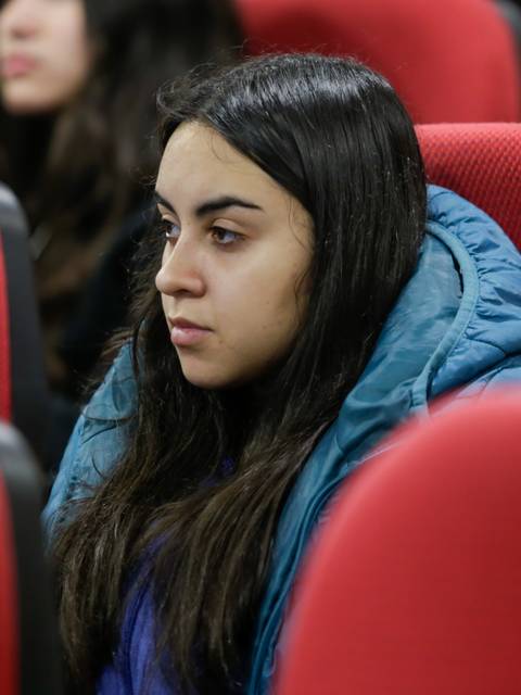 Una joven con chaqueta azul observa atentamente en una sala llena de asientos rojos.
