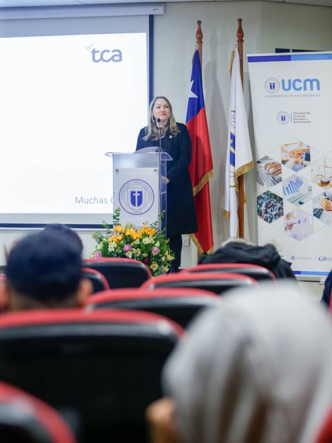 Una mujer habla en un podio durante un evento académico en un salón con banderas y un proyector al fondo.