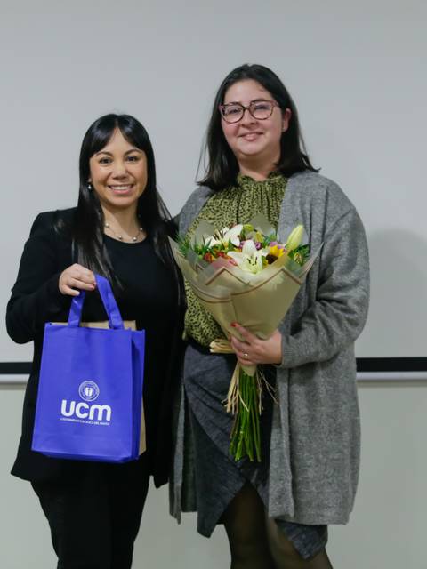 Dos mujeres posan sonrientes en un evento, una sosteniendo un ramo de flores y la otra una bolsa con el logo de UCM.
