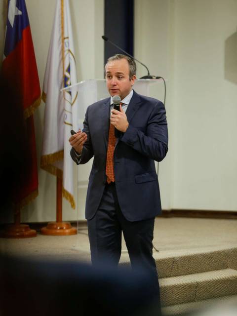 Un hombre de pie, vestido con un traje oscuro, habla al público mientras sostiene un micrófono en un evento.
