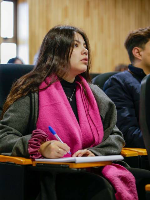 Una joven con una bufanda rosa toma notas en un aula.