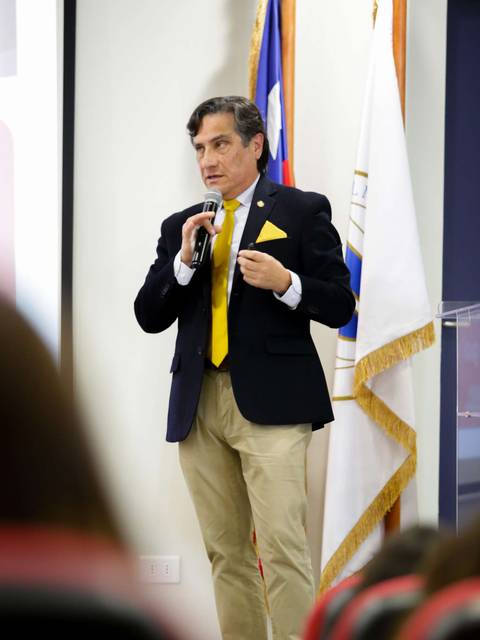 Un hombre hablando en una conferencia con un micrófono en mano, vestido con un traje oscuro y una corbata amarilla.
