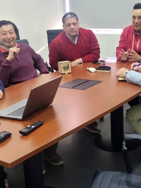 Un grupo de seis hombres se encuentra en una reunión en una oficina, conversando y sonriendo alrededor de una mesa.