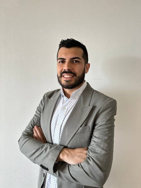 Un hombre con chaqueta gris y camisa blanca sonríe mientras cruza los brazos.