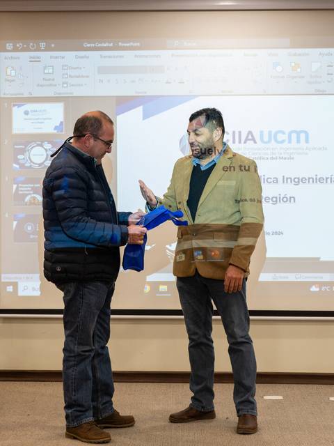 Dos hombres en un evento formal se intercambian un objeto frente a una presentación en pantalla.