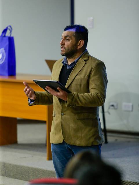 Un hombre con chaqueta clara habla frente a una audiencia en un salón de clases.