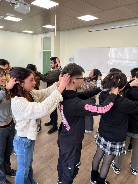 Un grupo de personas está realizando una actividad en un aula, con algunos de ellos tocándose las cabezas mientras otros observan.