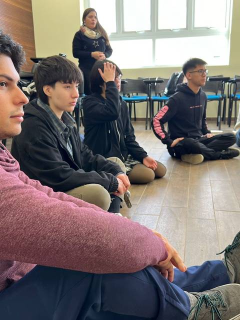 Un grupo de jóvenes sentados en el suelo, atentos a una persona que se encuentra de pie en el fondo de la sala.