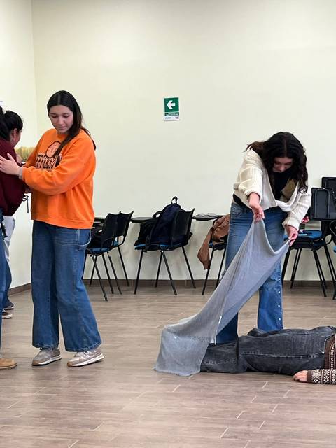 Un grupo de jóvenes actúan en una sala, con una persona en el suelo y otros interactuando entre sí.