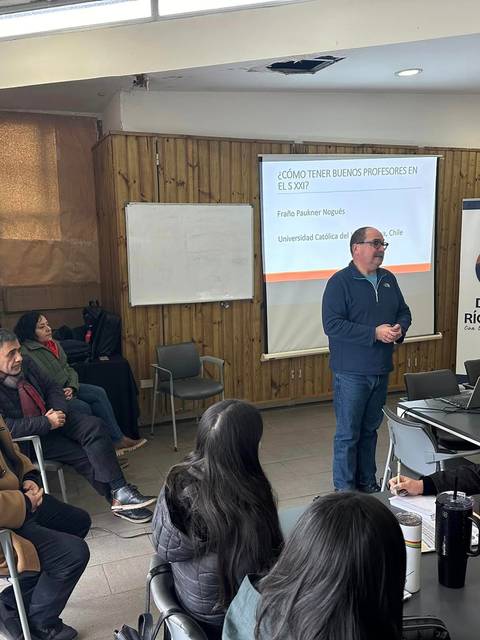 Una reunión en un aula donde un hombre de pie presenta un tema a un grupo de personas sentadas.