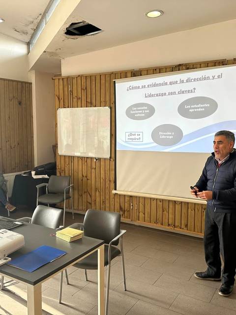 Un hombre está presentando en una sala con una presentación proyectada en una pantalla y un grupo de personas sentadas al fondo.