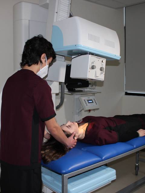 Un técnico de radiología está acomodando a una paciente sobre una mesa de examen frente a una máquina de rayos X.