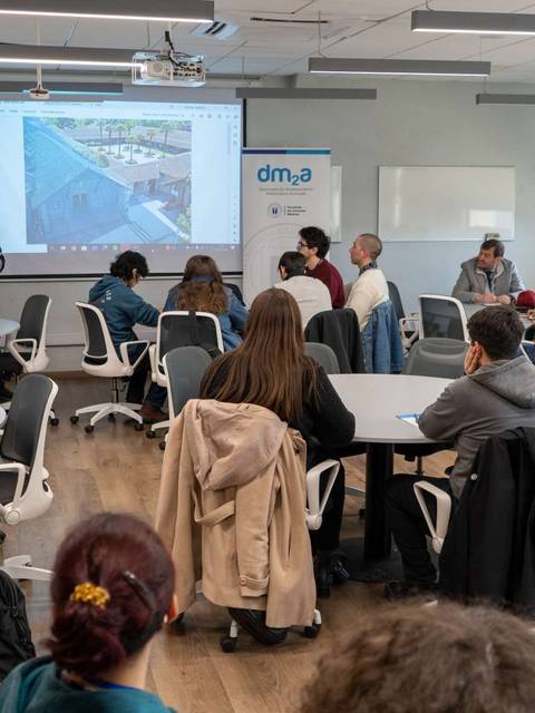 Un grupo de personas asiste a una presentación en un aula con pantallas y mesas redondas.