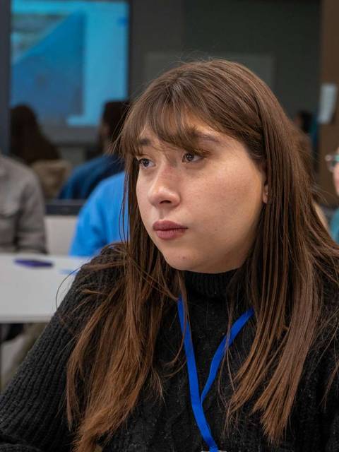 Un grupo de personas se sienta en una sala de reuniones, mientras una mujer con suéter negro y cabello largo mira pensativa hacia un lado.
