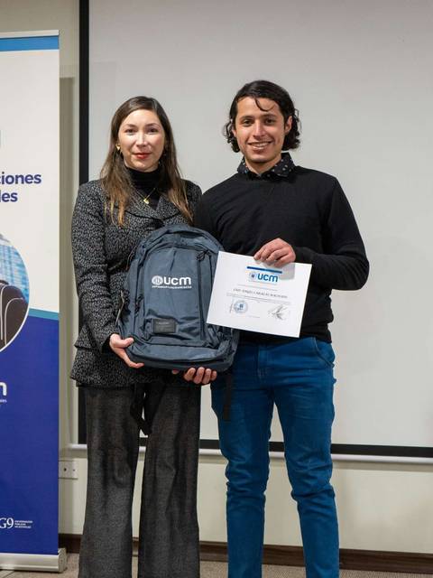 Dos personas posan en un evento académico, una de ellas sostiene un diploma y una mochila.