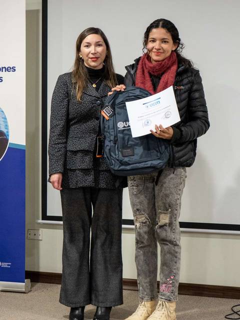 Dos mujeres posan juntas en un evento, una de ellas sostiene un certificado y una mochila.