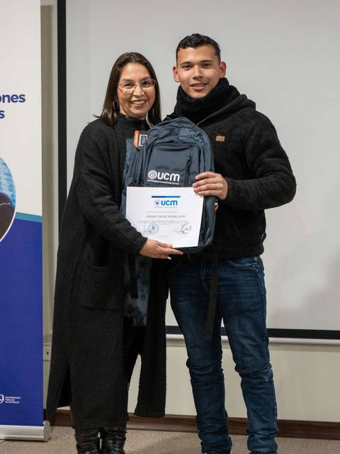 Dos personas posan juntas, una de ellas sosteniendo un certificado y una mochila con un logo.