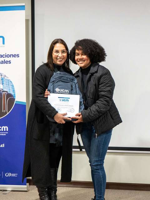 Dos mujeres posan juntas sosteniendo un diploma y una mochila en un evento formal.