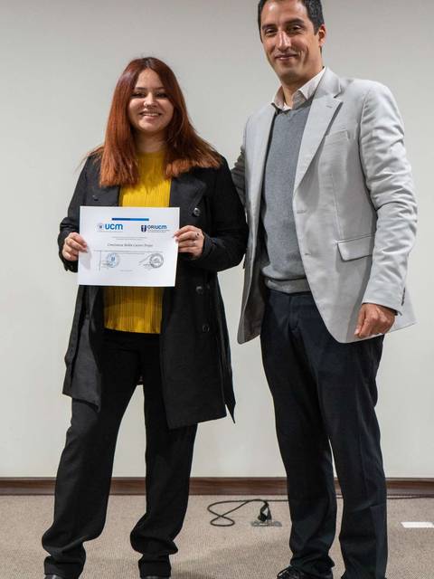 Una mujer y un hombre posan en un evento, ella sostiene un certificado mientras sonríen.