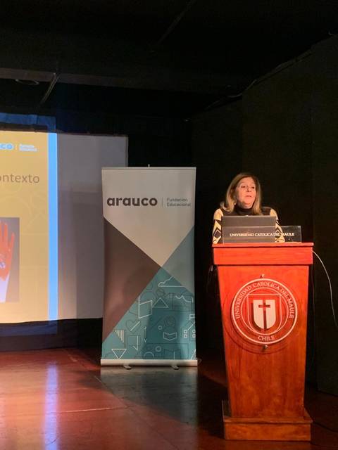 Una mujer está hablando en un escenario sobre la autoestima en el contexto escolar, mientras se proyecta una imagen colorida de manos en la pantalla detrás de ella.