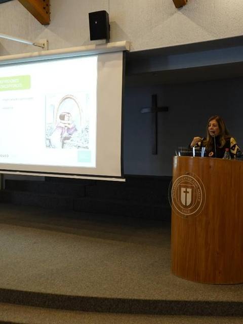 Una mujer está presentando en un evento en un auditorio con banderas al fondo.