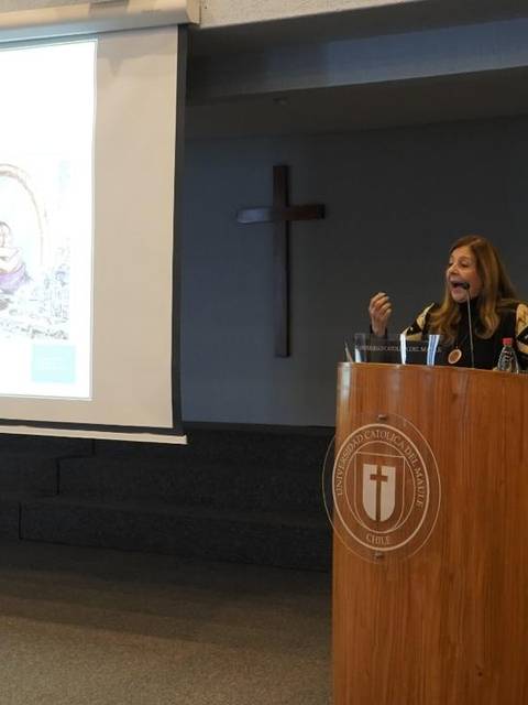 Una mujer habla frente a una presentación en una sala con bandera y un símbolo religioso.