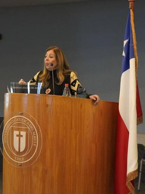 Una mujer está hablando en un podio junto a banderas de Chile y de la Universidad Católica.