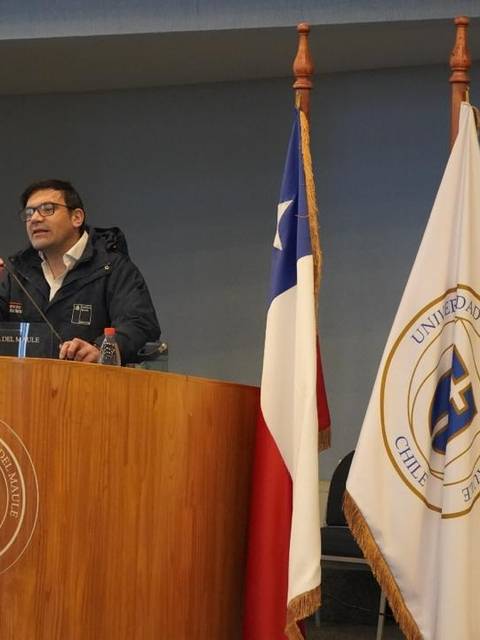 Un hombre está dando una charla en una universidad chilena, junto a banderas de Chile y de la universidad.