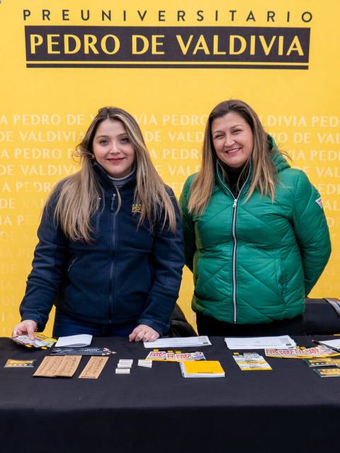 Dos mujeres sonrientes posando detrás de un stand con materiales promocionales de un preuniversitario.