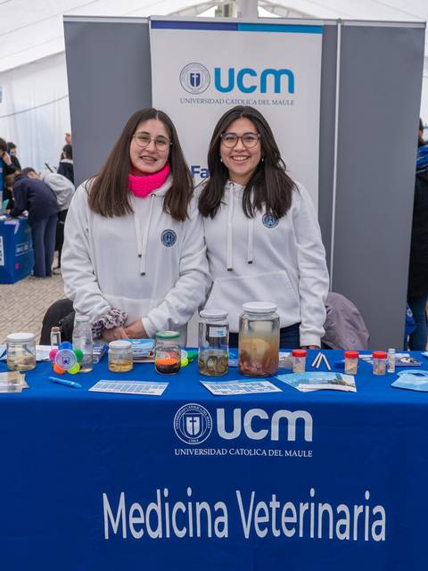 Dos estudiantes se encuentran en un stand de la Universidad Católica del Maule, presentando material relacionado con Medicina Veterinaria.