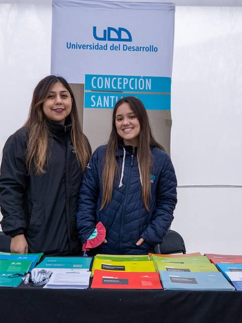 Dos chicas sonrientes están de pie detrás de una mesa con folletos de la Universidad del Desarrollo.
