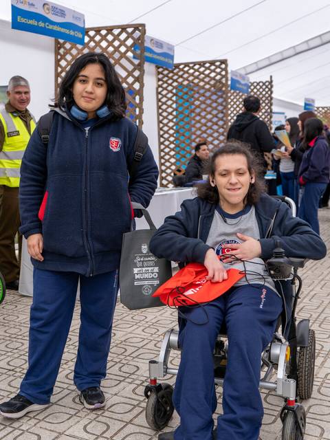 Dos jóvenes sonrientes, una de pie y otra en silla de ruedas, posan en un evento interior con varias personas y stands al fondo.