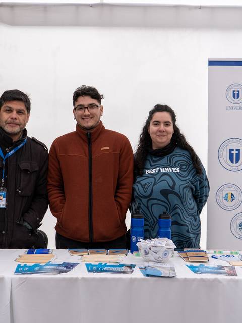 Tres personas posan frente a un stand de la universidad con información sobre la facultad de ciencias básicas.