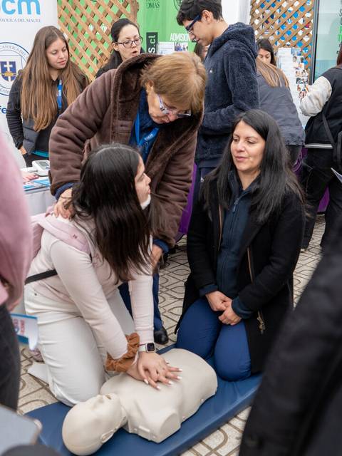 Una mujer está realizando maniobras de reanimación cardiopulmonar en un maniquí mientras otras personas observan en un evento educativo.