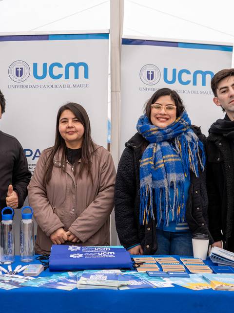 Cuatro jóvenes posan juntos frente a una mesa con productos promocionales de la Universidad Católica del Maule.