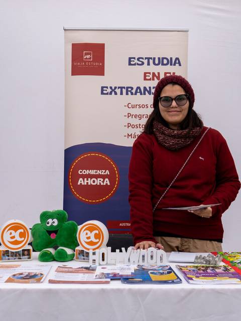 Una mujer sonriente presenta información sobre estudios en el extranjero en una feria educativa.