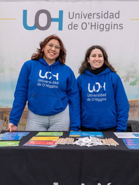 Dos mujeres sonrientes están de pie frente a una mesa con material promocional de la Universidad de O'Higgins.